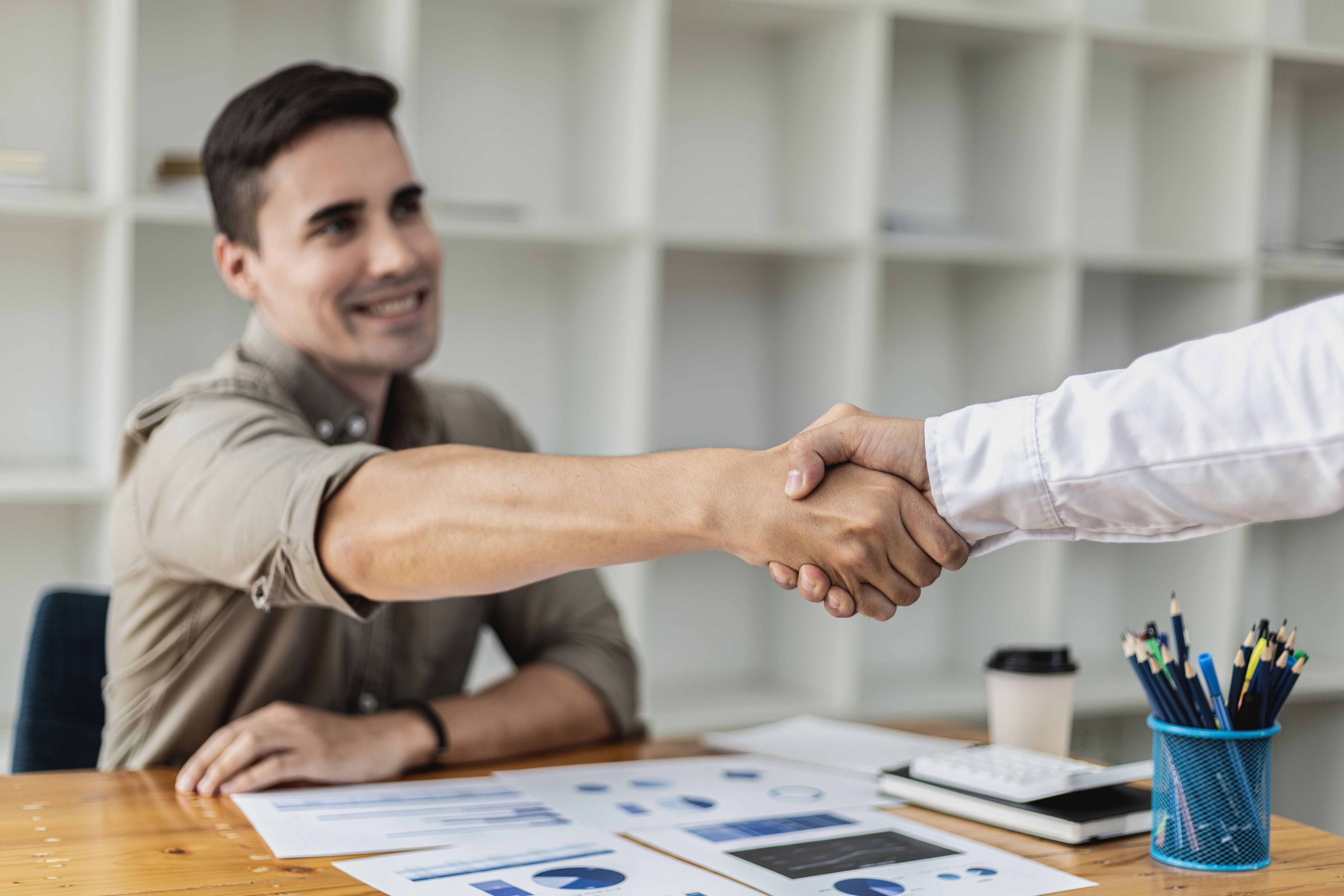 Ein Mann sitz am Schreibtisch und streckt seine Hand zum Händedruck entgegen. Seine Hand wird von einem zweiten Mann außerhalb des Bildes geschüttelt. 
