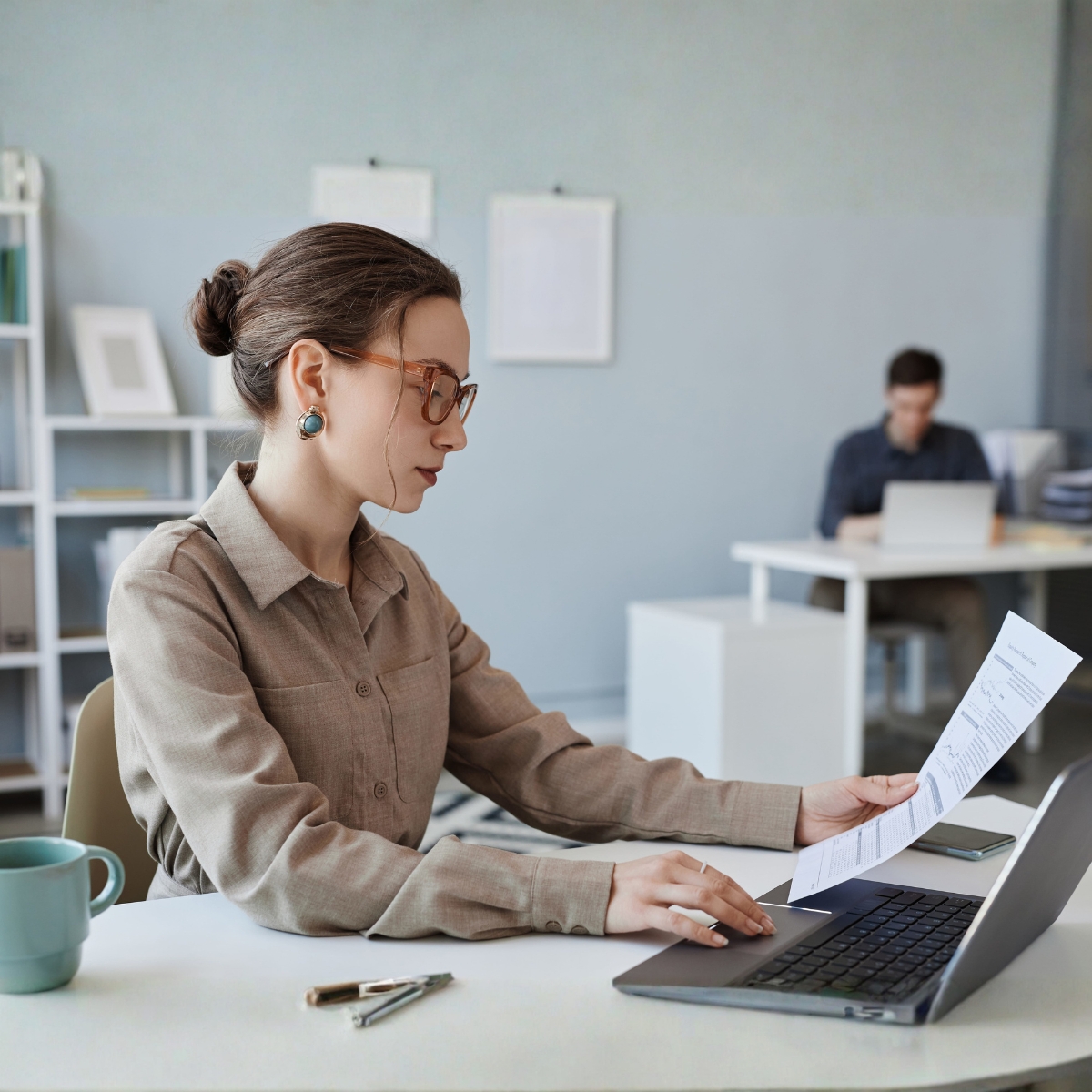 Eine Frau hat ein Dokument in der Hand und bearbeitet es am Computer; im Hintergrund arbeitet ein Mann am Laptop.