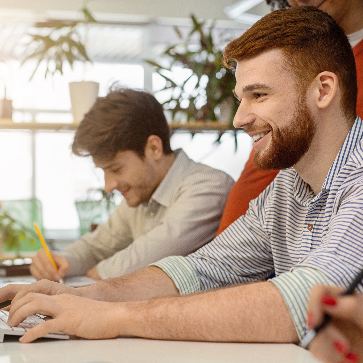 Zwei Männer arbeiten an Laptops. Der Mann im Vordergrund lächelt. Hinter im steht eine weitere Person.