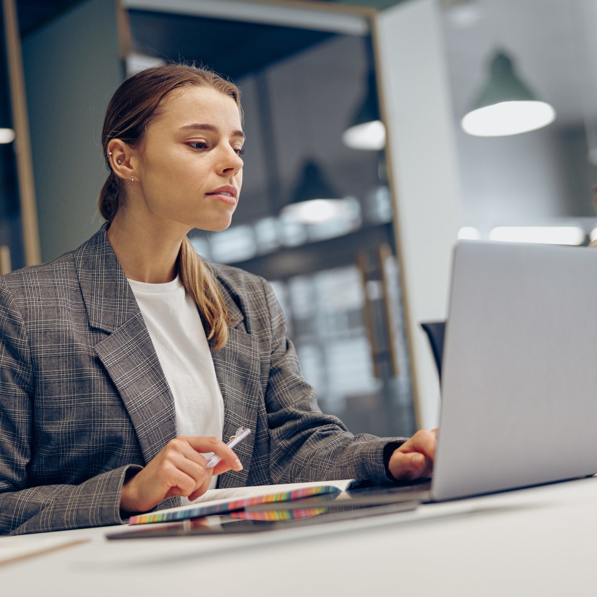 Eine Frau sitzt vor einem Laptop und blickt konzertiert auf den Bildschirm. Sie hat einen Stift in der Hand. 