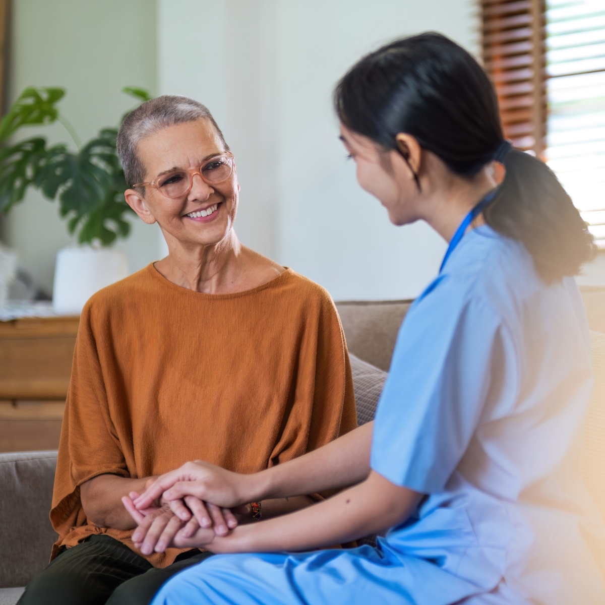 Eine Pflegerin sitzt mit einer Patientin auf einem Sofa. Die Pflegerin hält behutsam die Hände der Patientin.