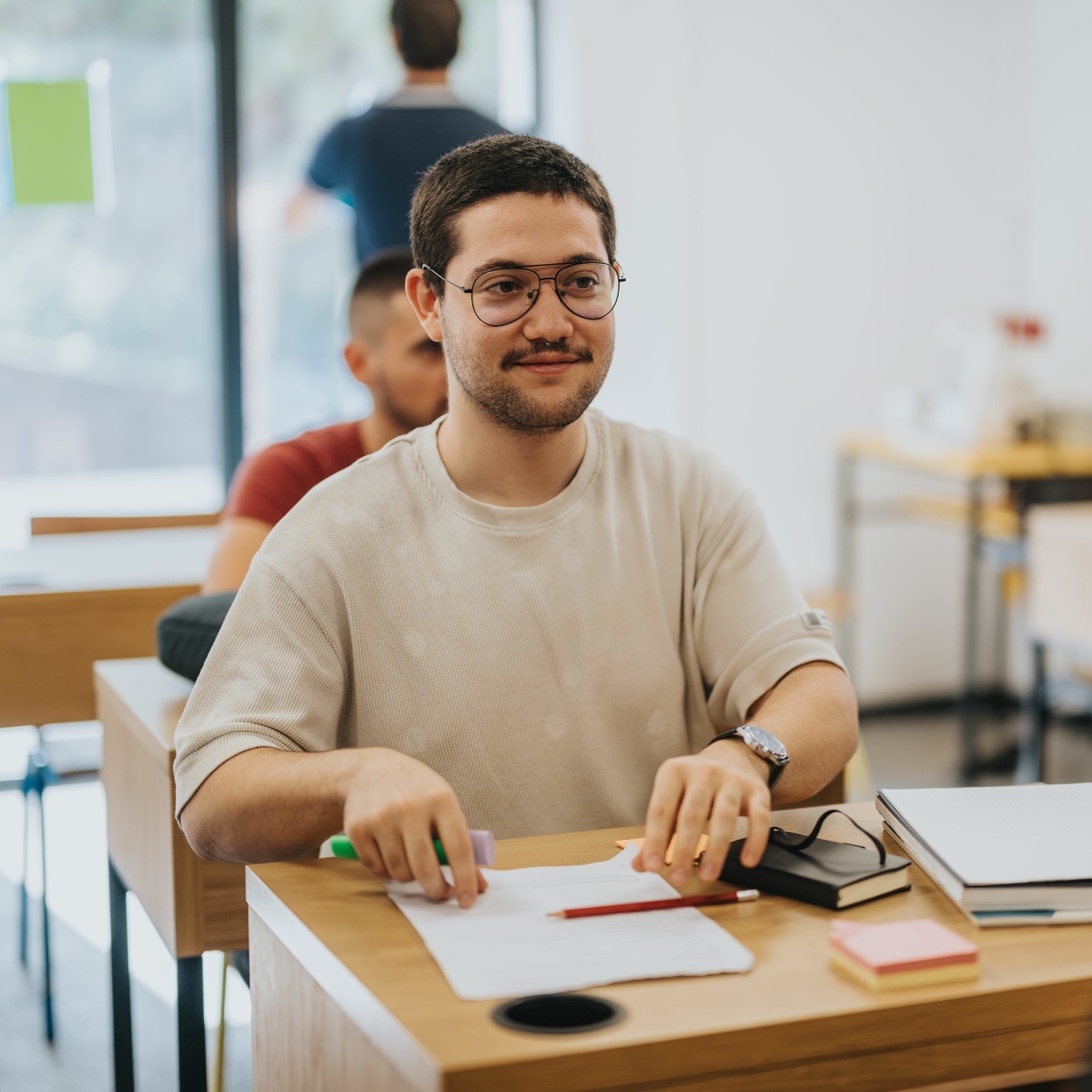 Mehrere Junge Männer sind in einem Klassenzimmer. Der Mann im Vordergrund hat auf seinem Tisch Arbeitsmaterial liegen.